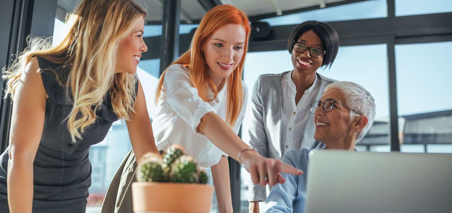 Eine vielfältige Gruppe von vier Frauen arbeitet in einem hellen Büro gemeinsam an einem Laptop. Eine zeigt auf den Bildschirm, während die anderen lächeln – ein Ausdruck von Teamarbeit und positiver Zusammenarbeit.