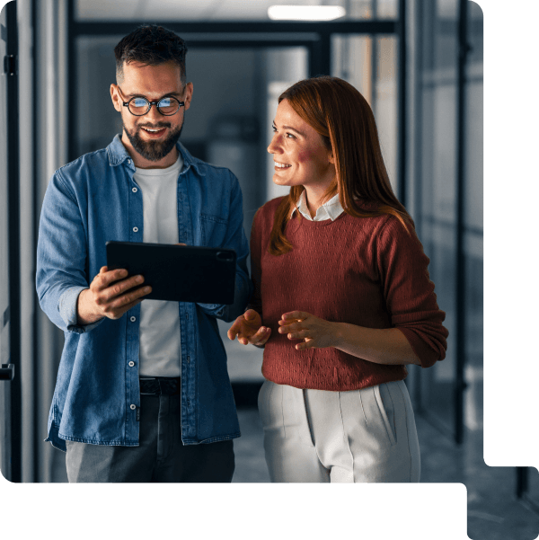 Zwei Personen stehen in einem modernen Büroflur und betrachten gemeinsam ein Tablet. Die Personen tragen Business-Casual-Kleidung und befinden sich vor einer Glaswand mit unscharfem Hintergrund. Die Szene vermittelt digitale Zusammenarbeit und den gemeinsamen Einsatz moderner Technologien im Arbeitsalltag.