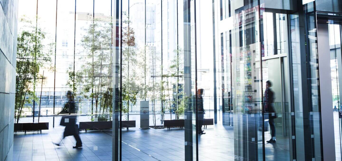 Photograph of a modern office building lobby with large glass walls and reflections creating a layered effect. Several blurred figures of people walking and standing are visible, emphasizing movement and activity within a sleek, urban environment.