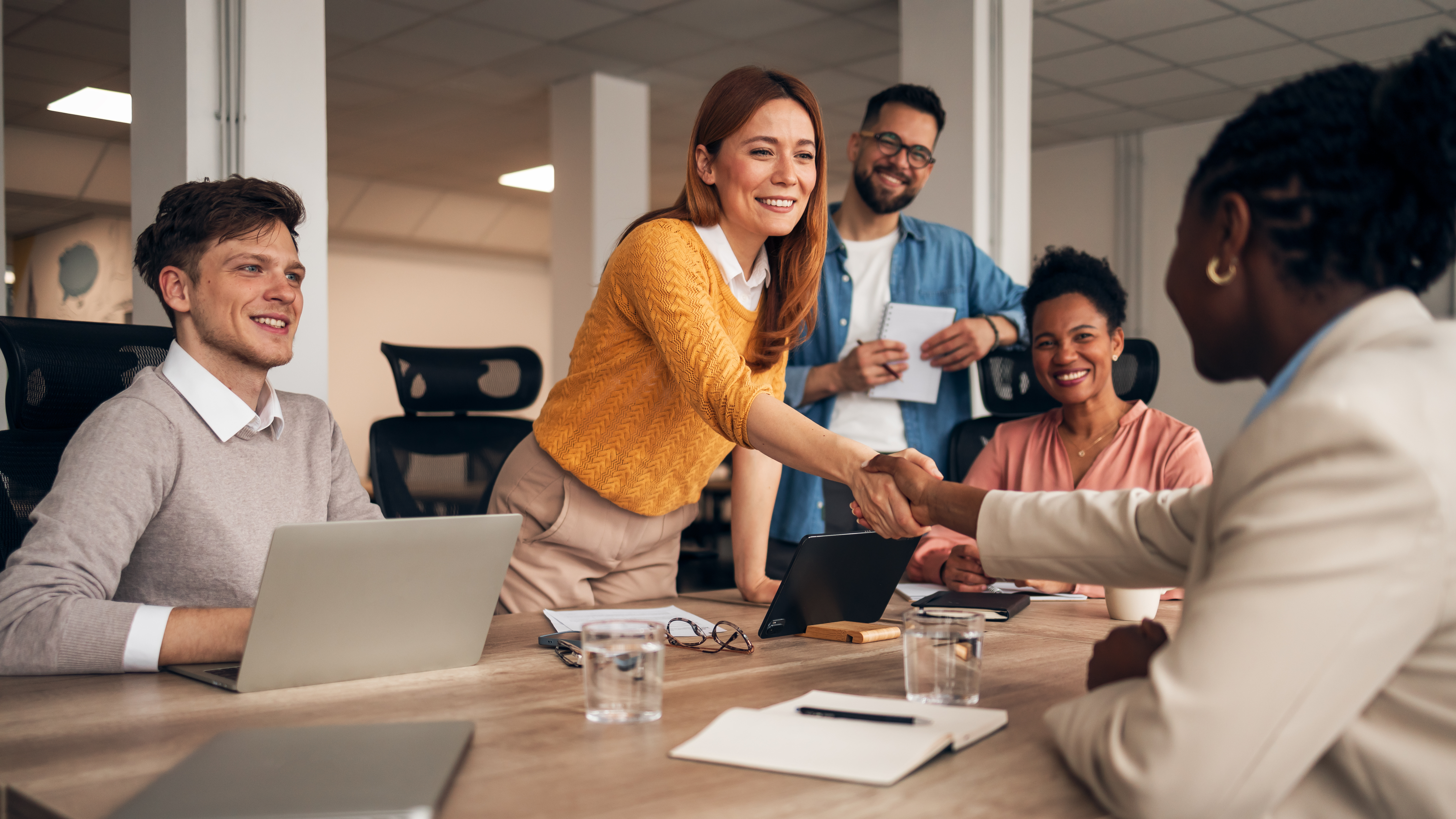 A diverse group in a meeting room, smiling as a woman in a yellow sweater shakes hands with a colleague across the table. A friendly, collaborative atmosphere.