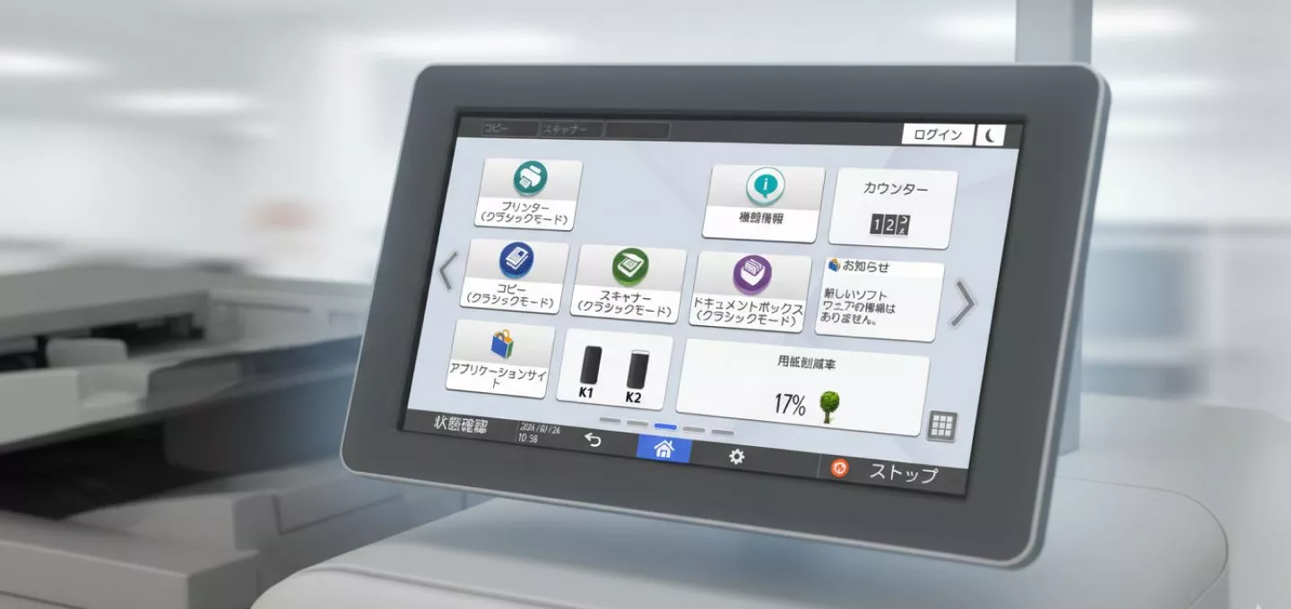 A close-up, angled view of a digital control panel on a Ricoh printer, with a user interface. The screen displays various icons and menus, including 